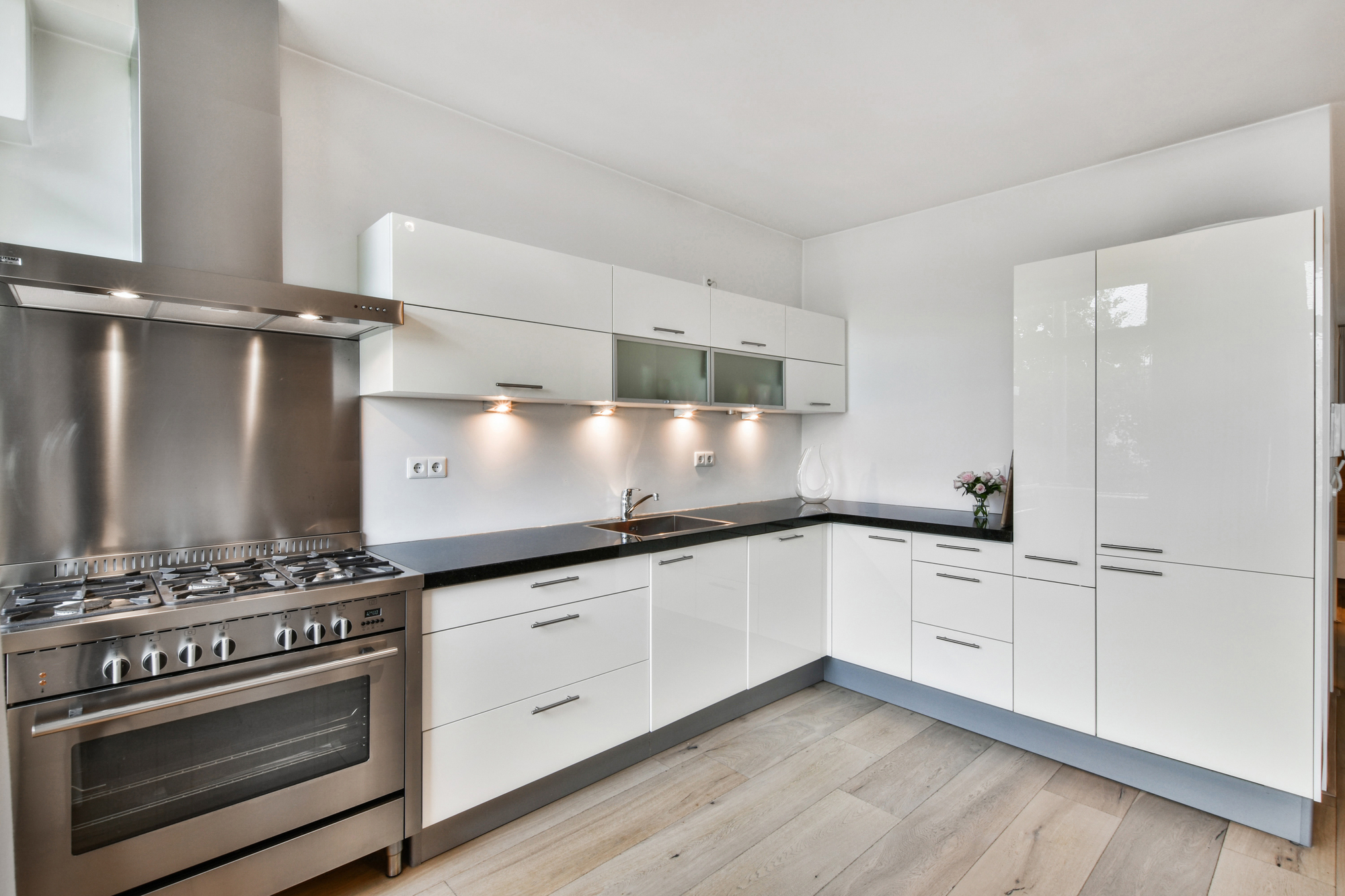 A luxury kitchen with white walls, white drawers, a white refrigerator, black countertops with a sink, and a steel stove and oven.