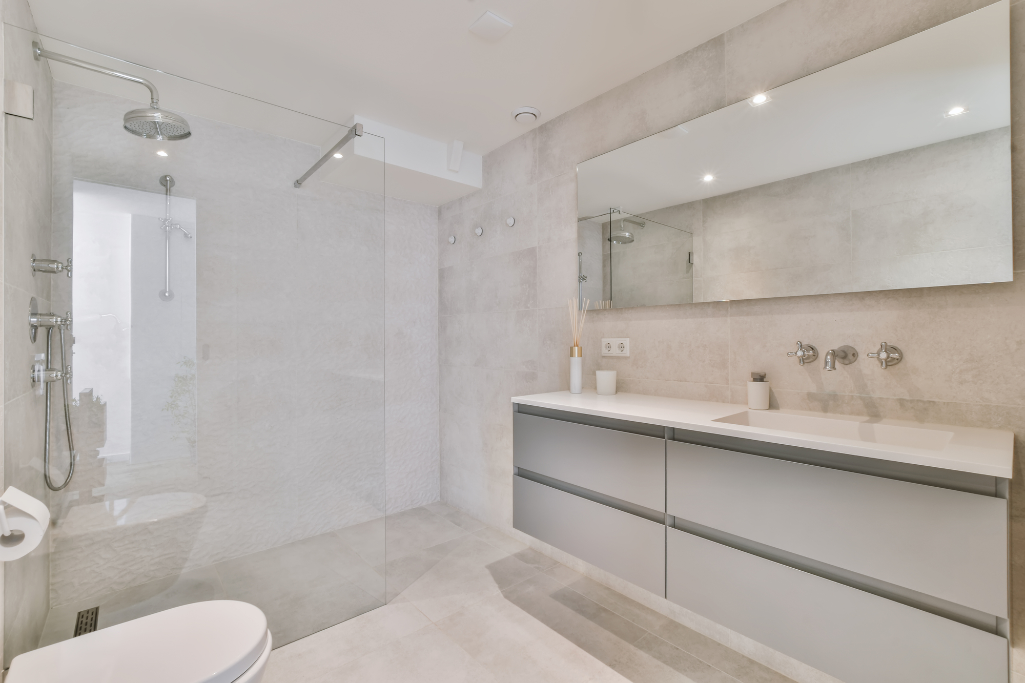 A remodeled bathroom with a walk-in shower and a  vanity with gray drawers and a wide mirror over it.