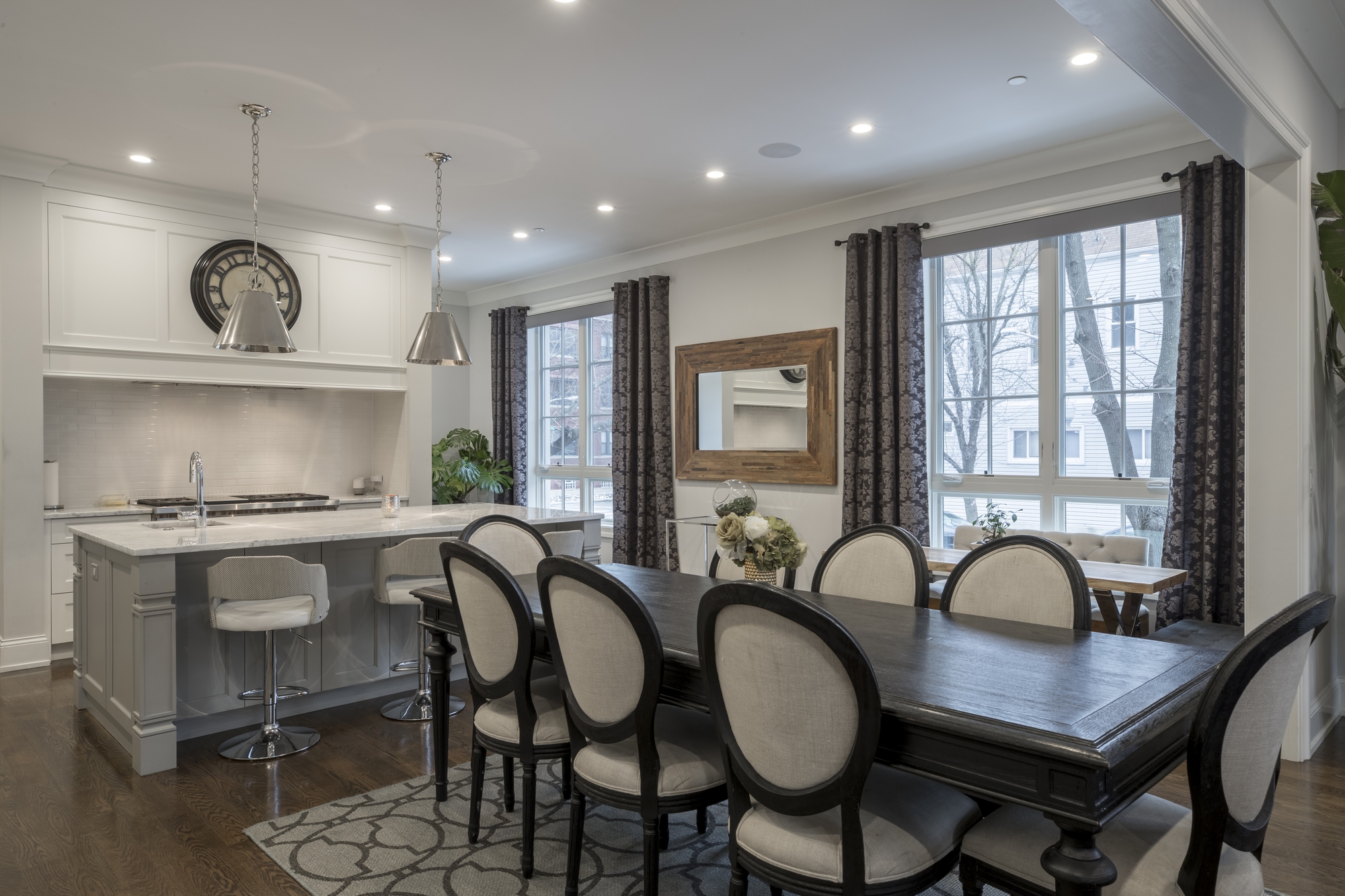 A dining room with a wood table and chairs, large windows, and a kitchen with marble countertops.