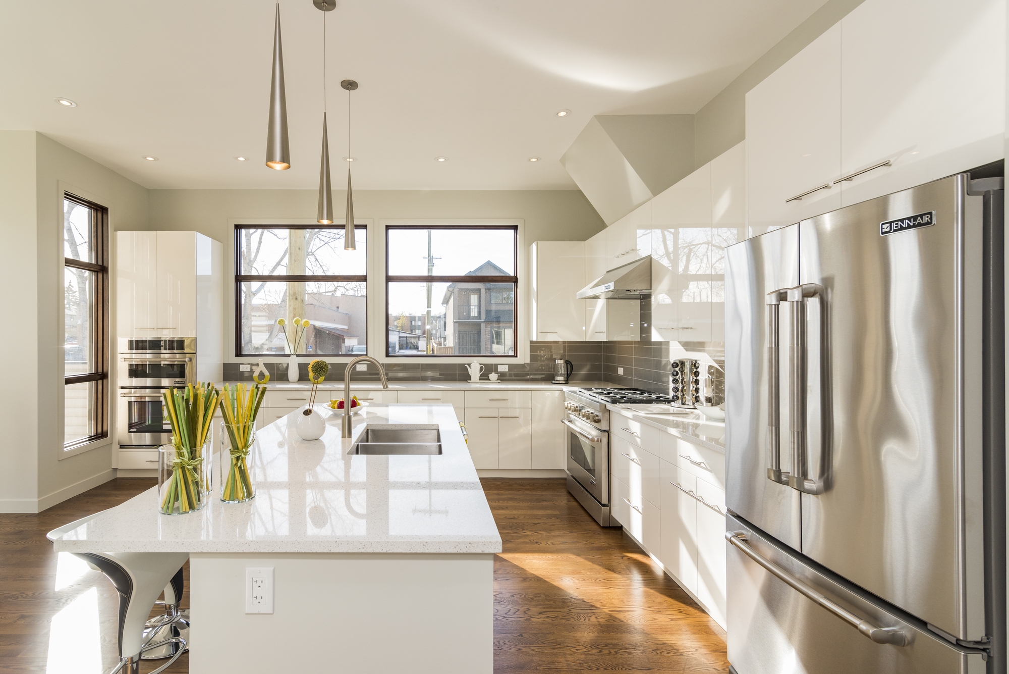 A modern kitchen with a white island, sink, a silver refrigerator and freezer, and large windows looking outside.