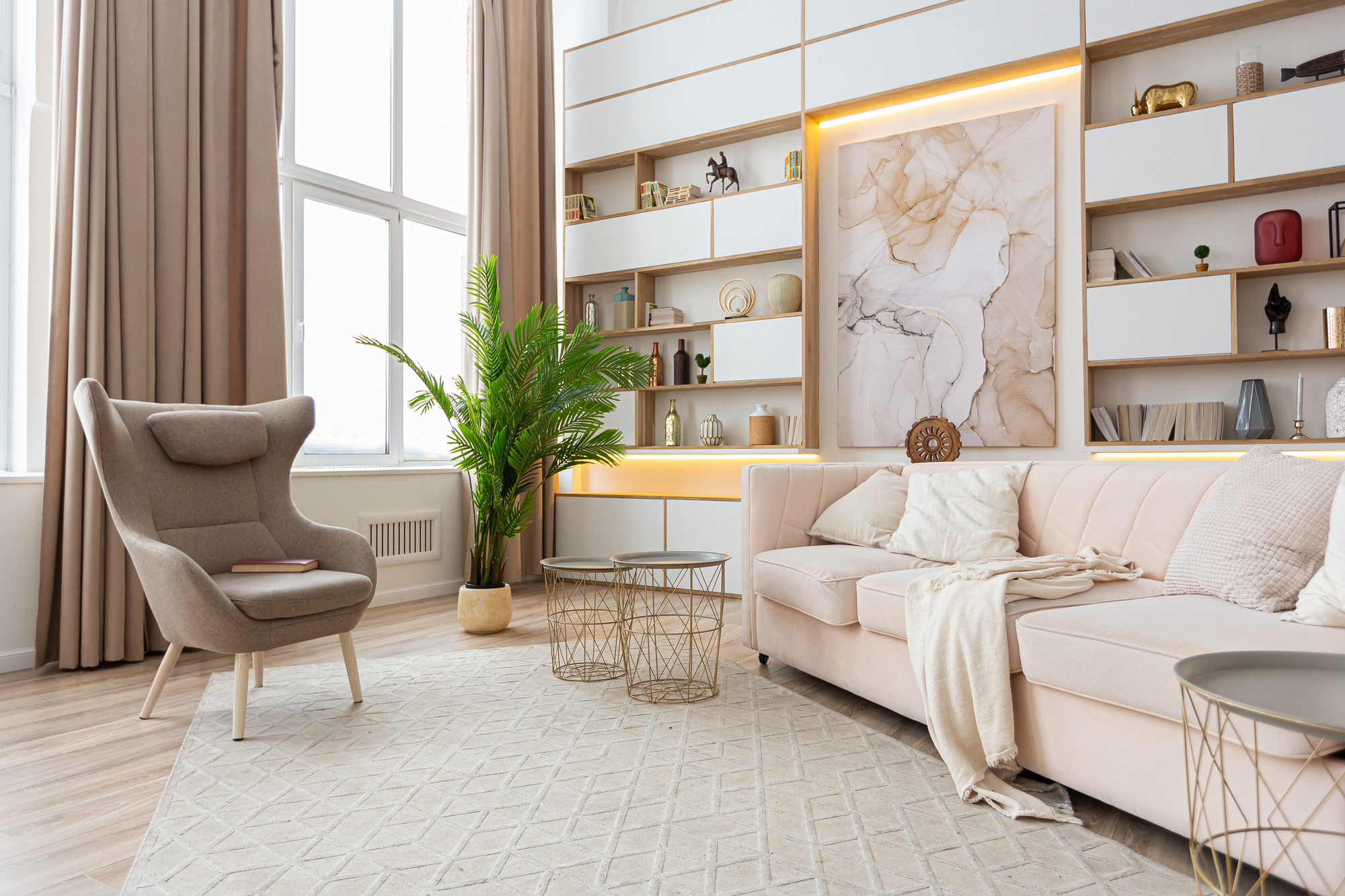 A luxury living room with an off-white couch, a gray chair, large windows, and a white wall with drawers and shelves.