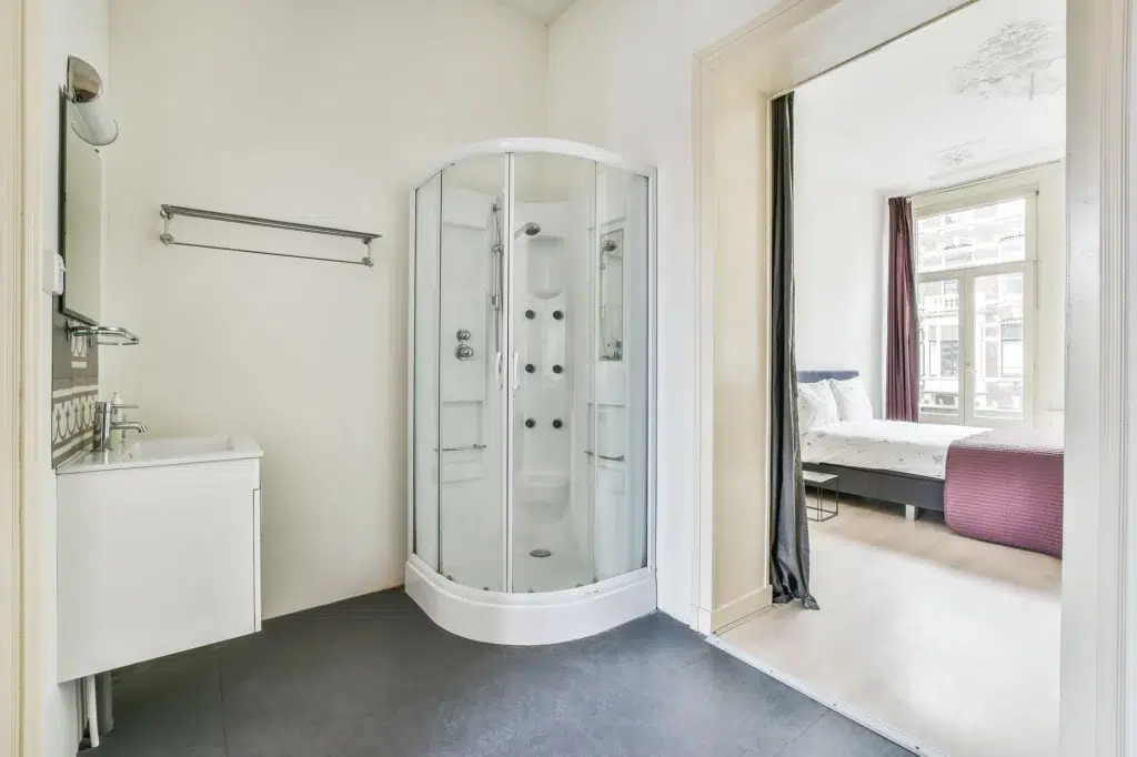 Walk-in acrylic shower in a bathroom with a sink looking out into a bedroom with a bed and window.