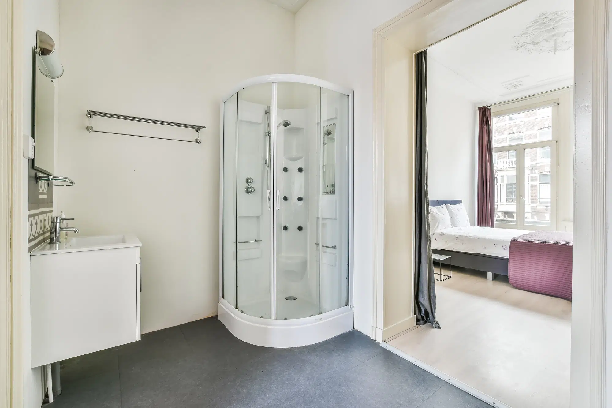 Walk-in acrylic shower in a bathroom with a sink looking out into a bedroom with a bed and window.
