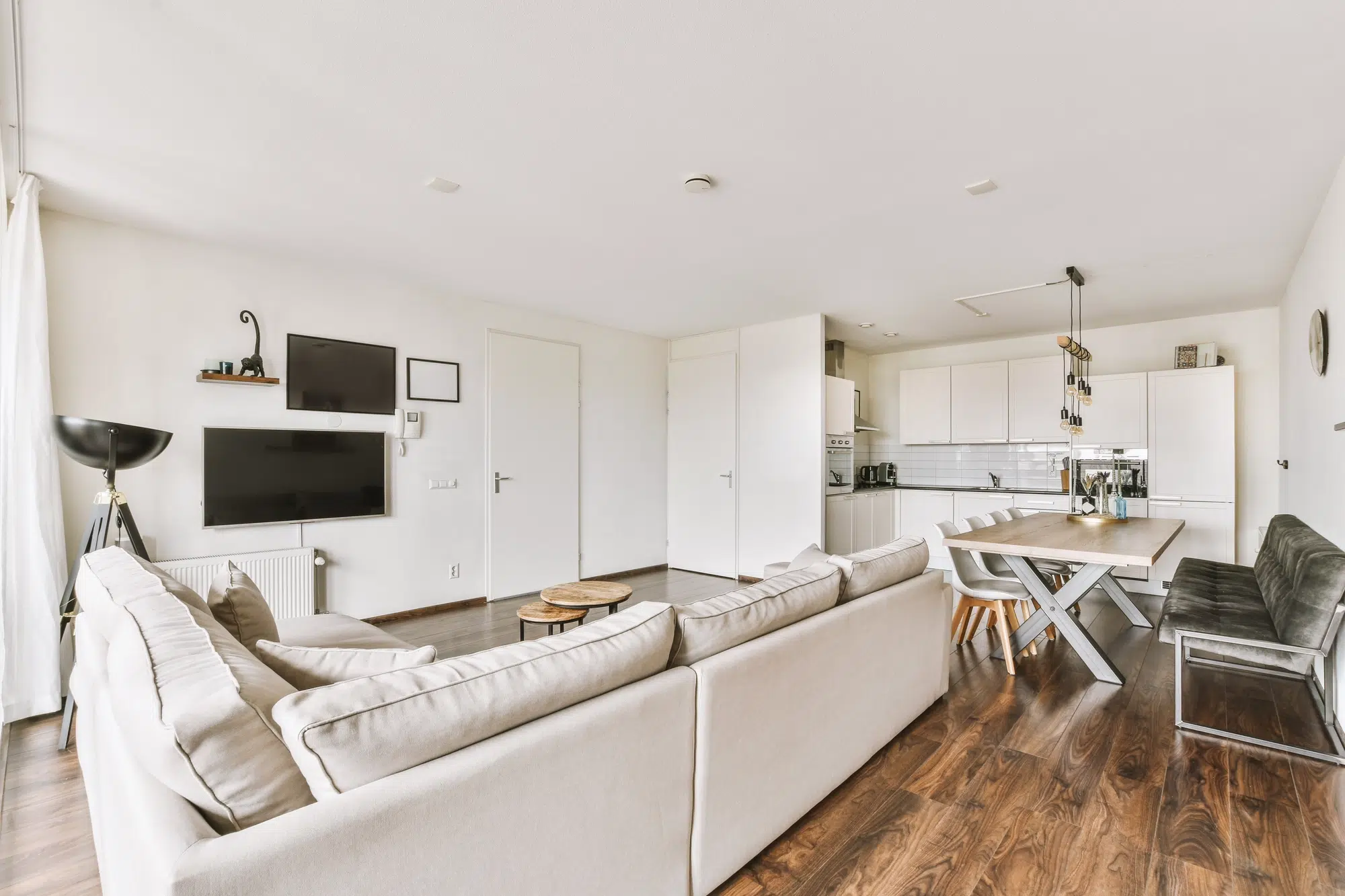 A spacious living room with a long white couch, white walls, a television on the wall, a dining table, and a kitchen with white cabinets.