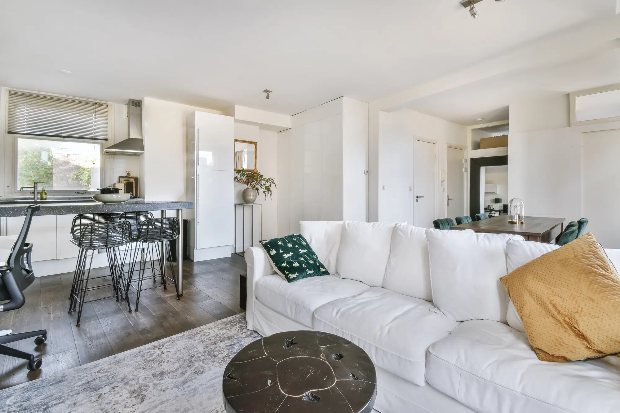 A living room with white walls, a white couch, a brown table, a dining table, and a kitchen in the background.