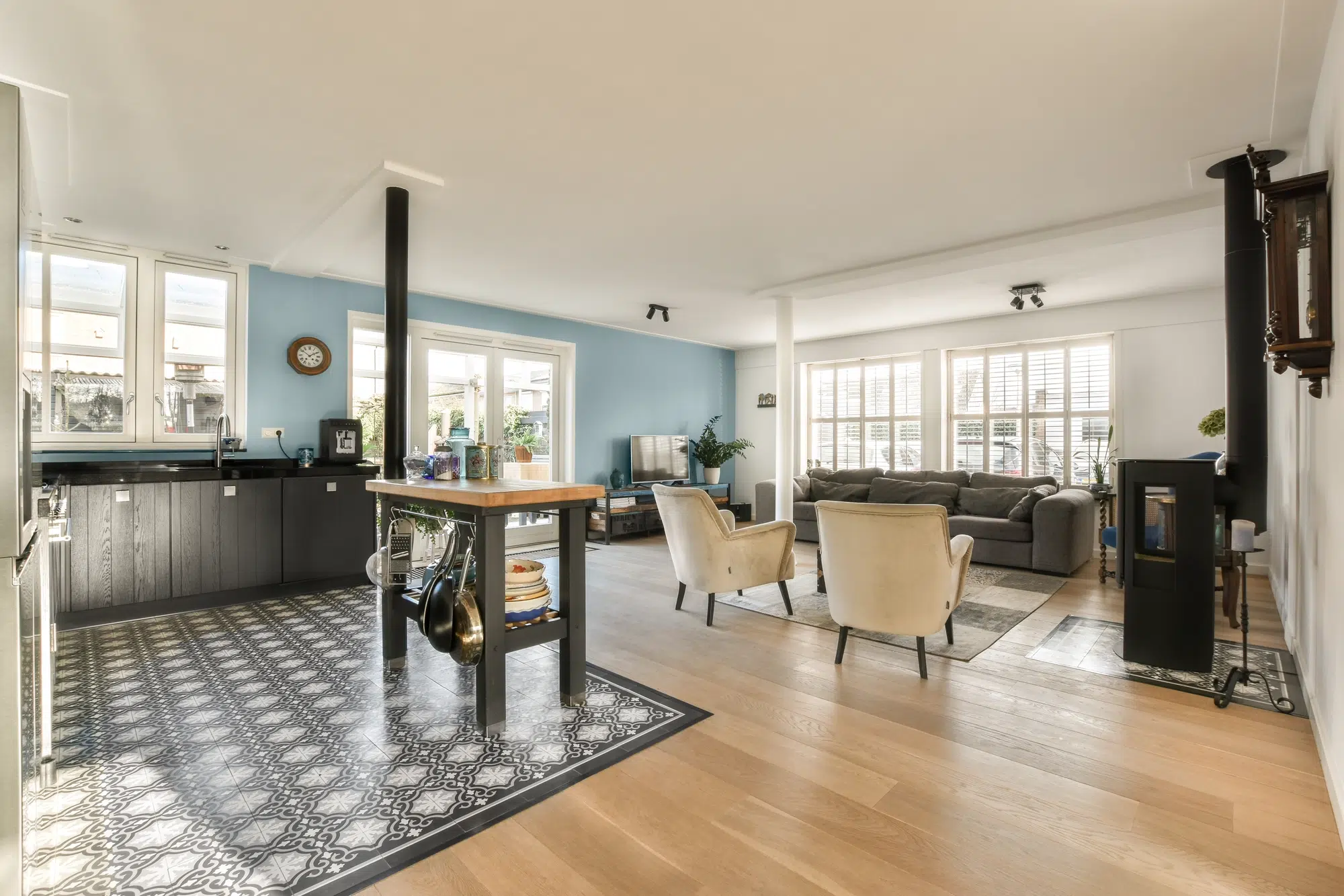 A kitchen and living room with hardwood floors, blue and white walls, two tan chairs, and a gray couch.