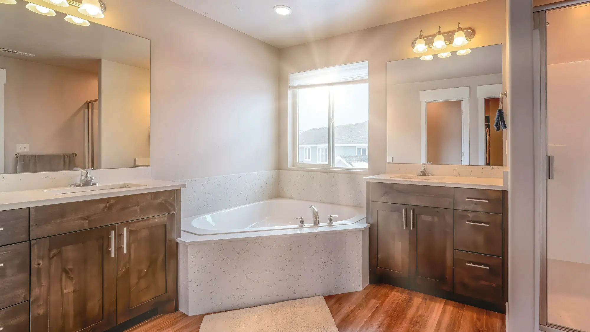 A bathroom with a white corner bathtub, two wooden vanities with mirrors and overhead lighting, and hardwood flooring.