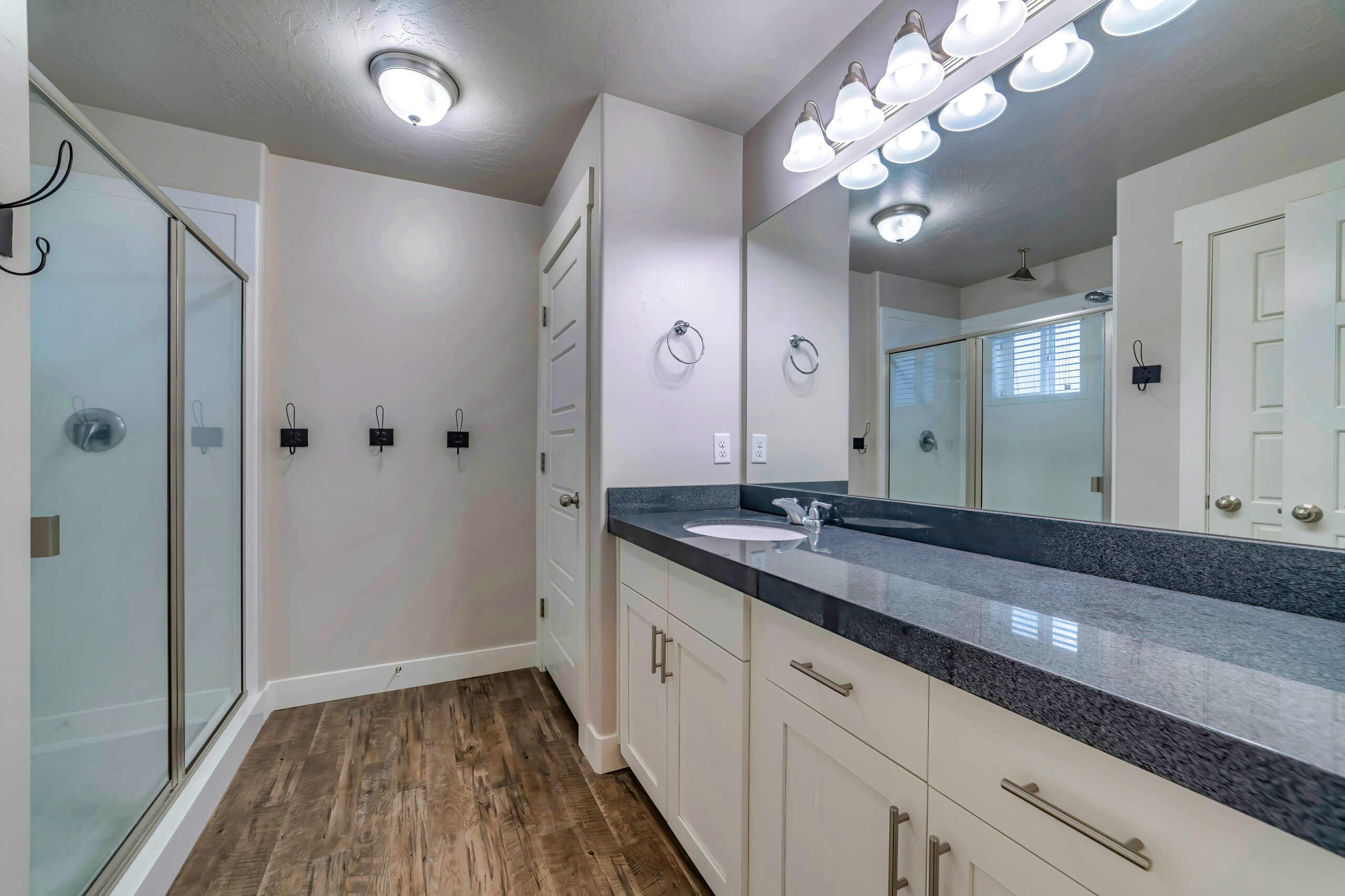 A narrow bathroom with hardwood floors, a blue countertop, white cabinets, and a walk-in shower with a glass door.