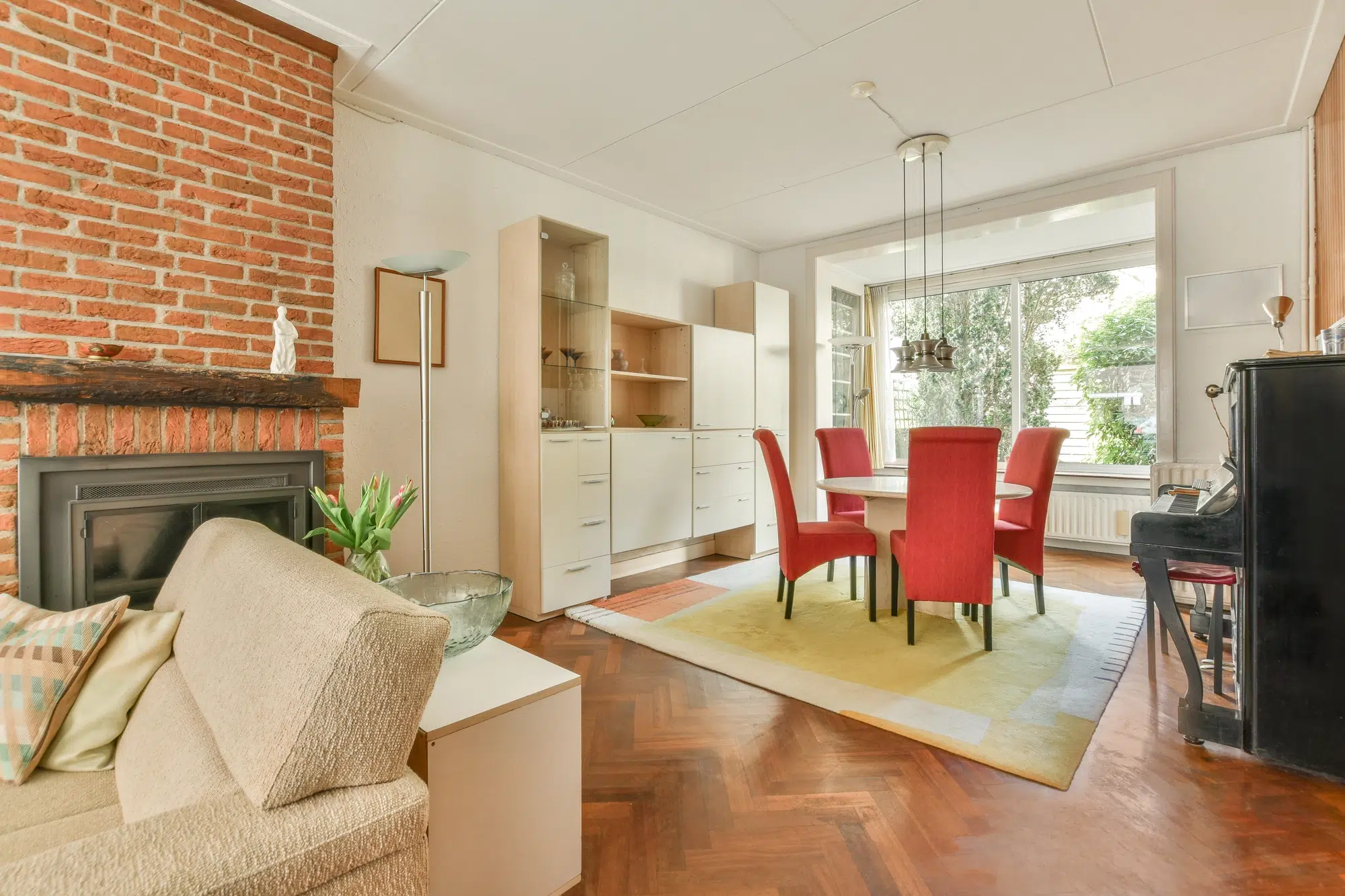 A tan living room with hardwood flooring, a tan couch, a brick fireplace, a table with chairs, and a black piano.