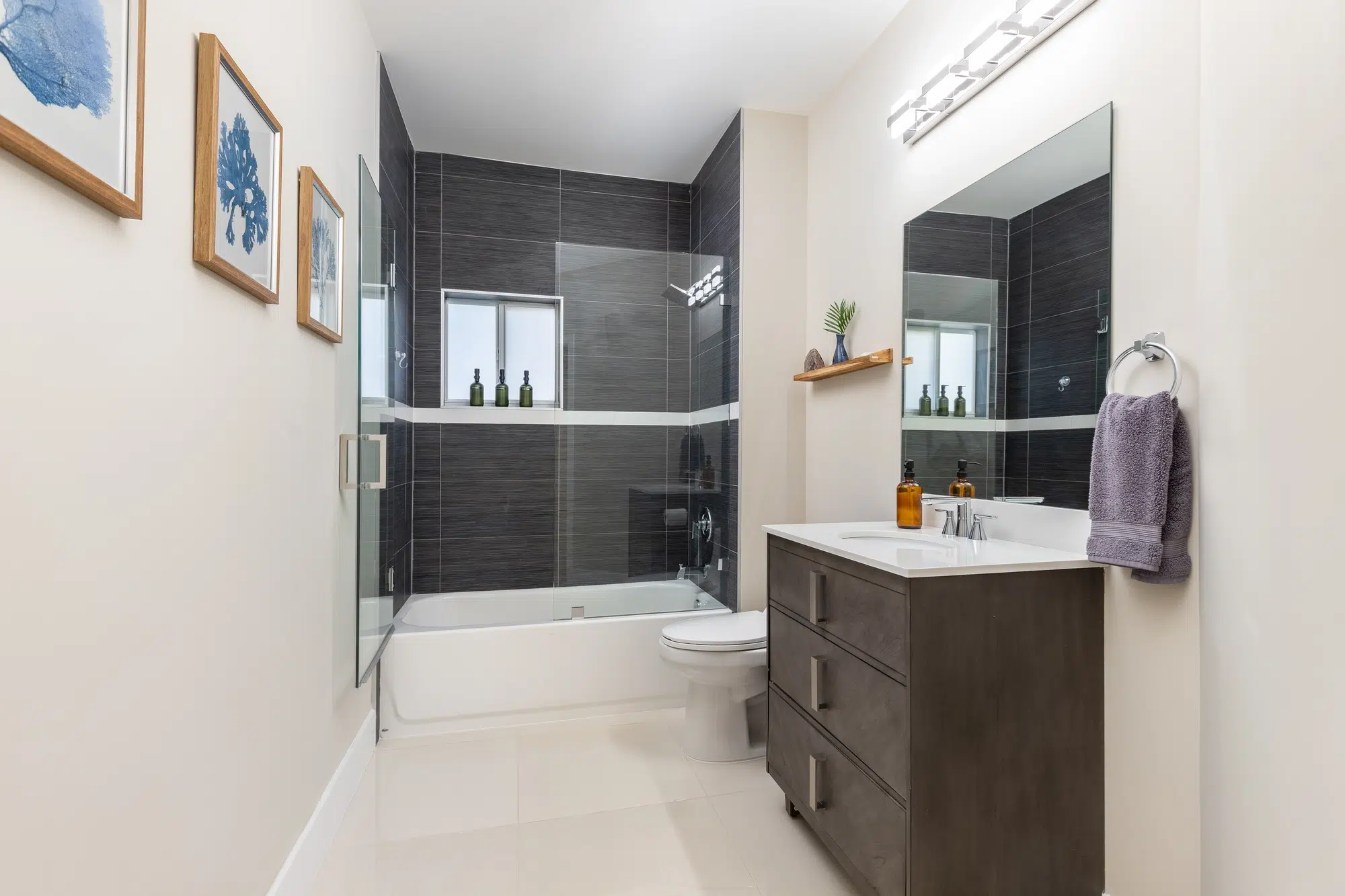 A bathroom with white walls, white tile floors, a wooden vanity, a white toilet, and a white bathtub with a glass door.