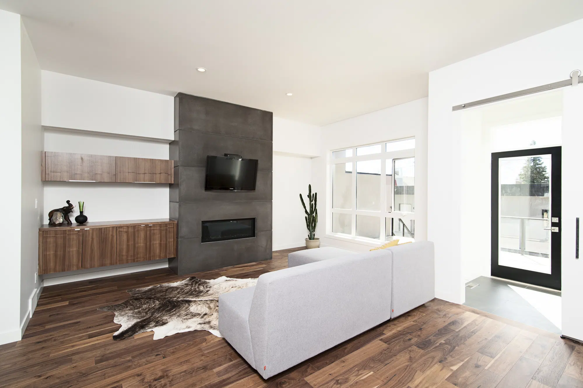 A living room with wooden flooring, a gray couch, and wood shelves with a television on the wall.