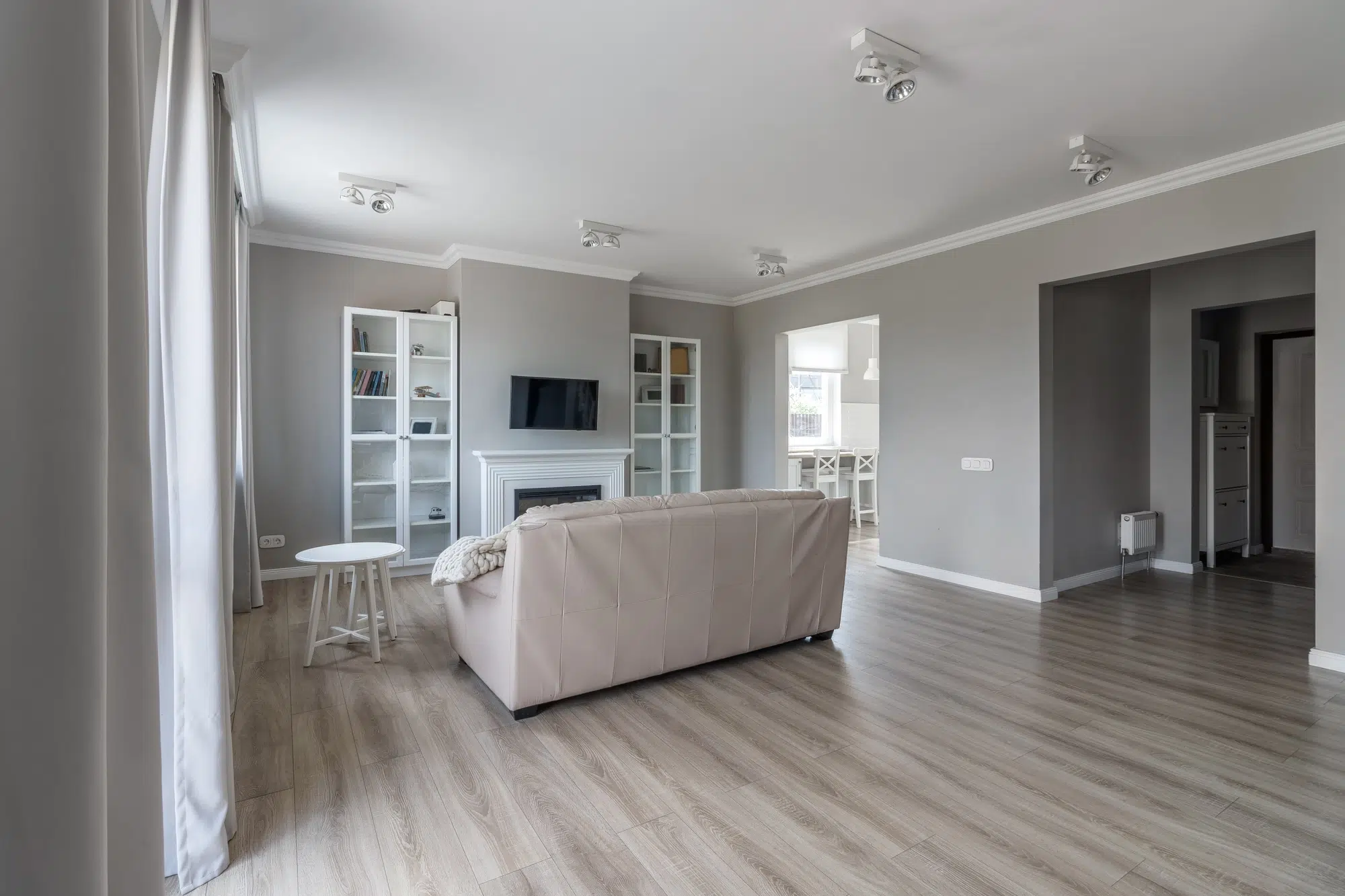 A living room with hardwood flooring, gray walls, a beige couch, a white table, and a television on the wall.