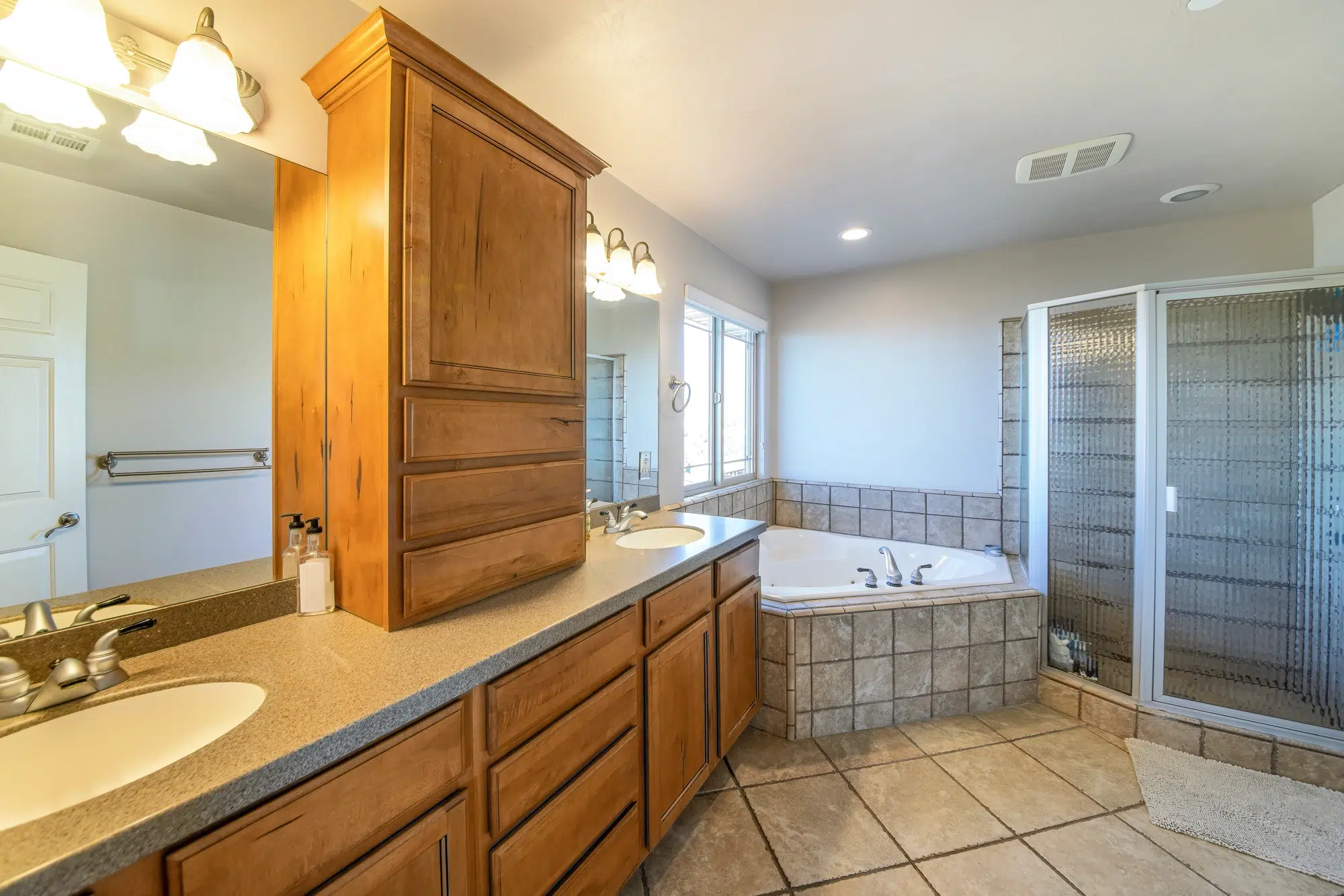 A bathroom with wood drawers, a marble countertop with two sinks, a white bathtub, and a walk-in shower with a glass door.