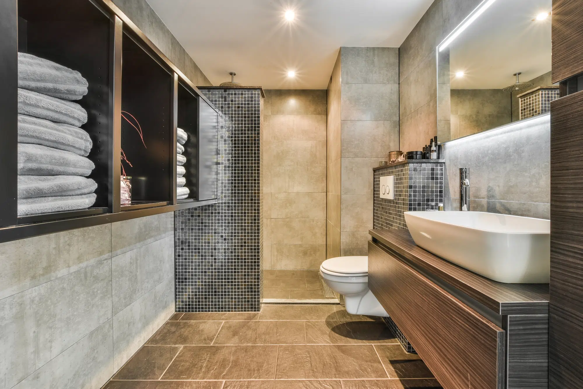 A bathroom with tile flooring, wooden cabinets for towels, a wooden vanity, a white toilet, and a walk-in shower.