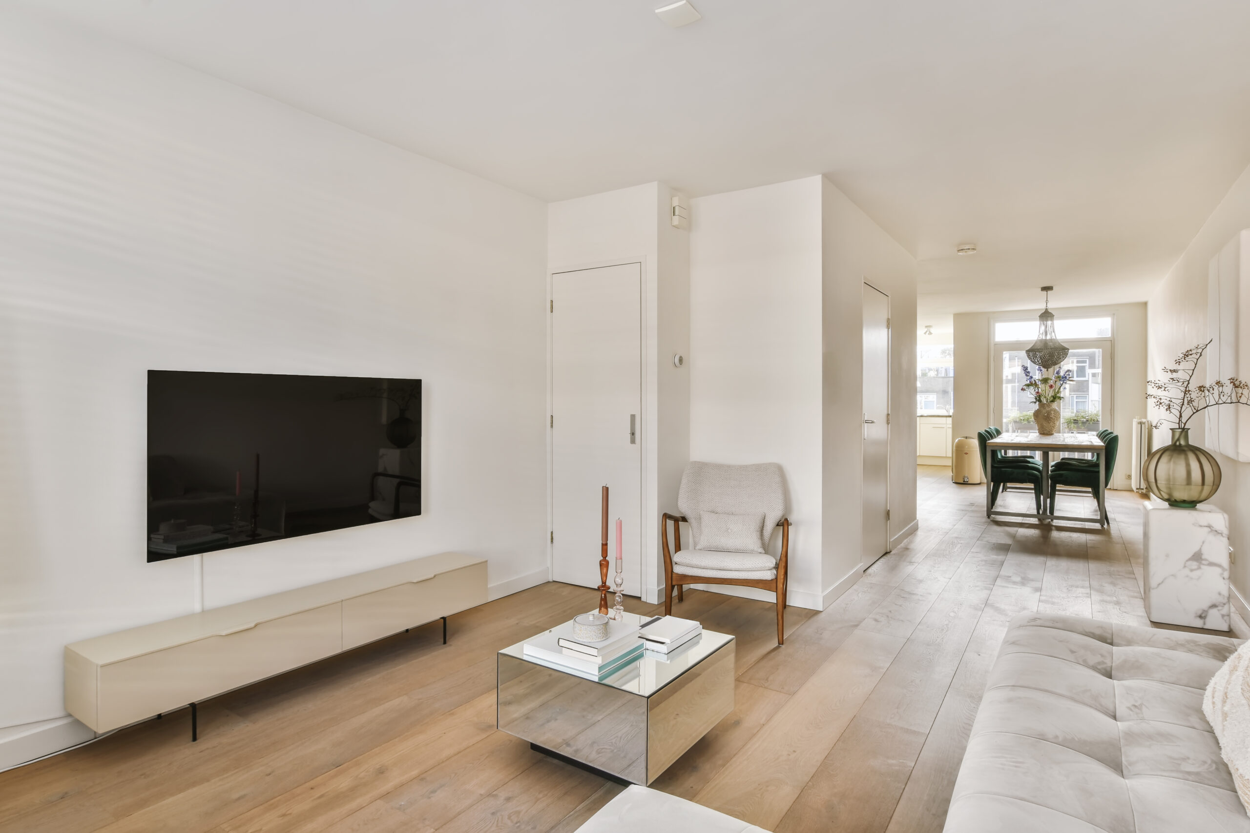A living room with hardwood flooring, a gray couch, a television on the wall, a silver reflective table, and a dining room in the background.