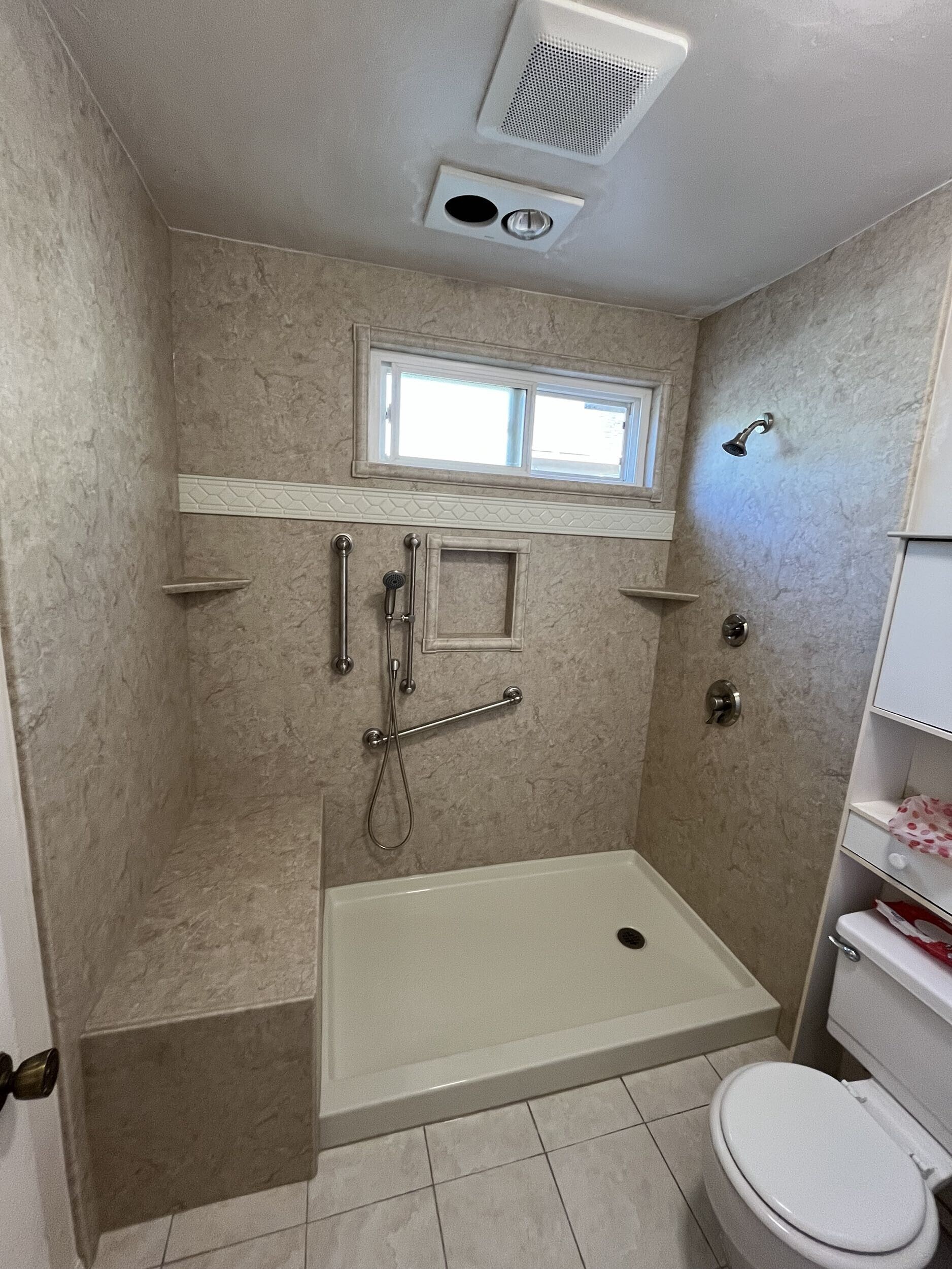A bathroom featuring a walk-in shower with beige marble-look wall panels, built-in corner shelves, multiple grab bars, and a handheld showerhead.