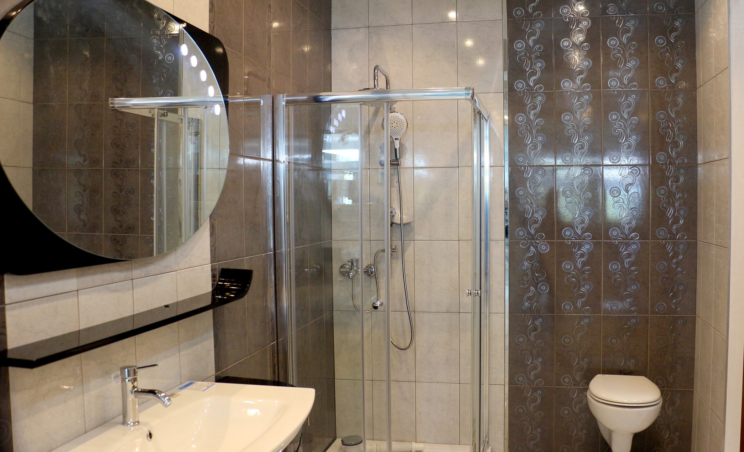 A modern bathroom featuring a glass-enclosed corner shower with chrome fixtures, glossy patterned accent tiles, a wall-mounted toilet, and a white vanity.
