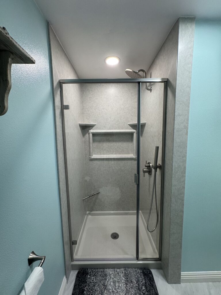 Modern walk-in shower with gray wall panels, a glass sliding door, a rainfall showerhead with handheld sprayer, a built-in shelf, and a white shower base.