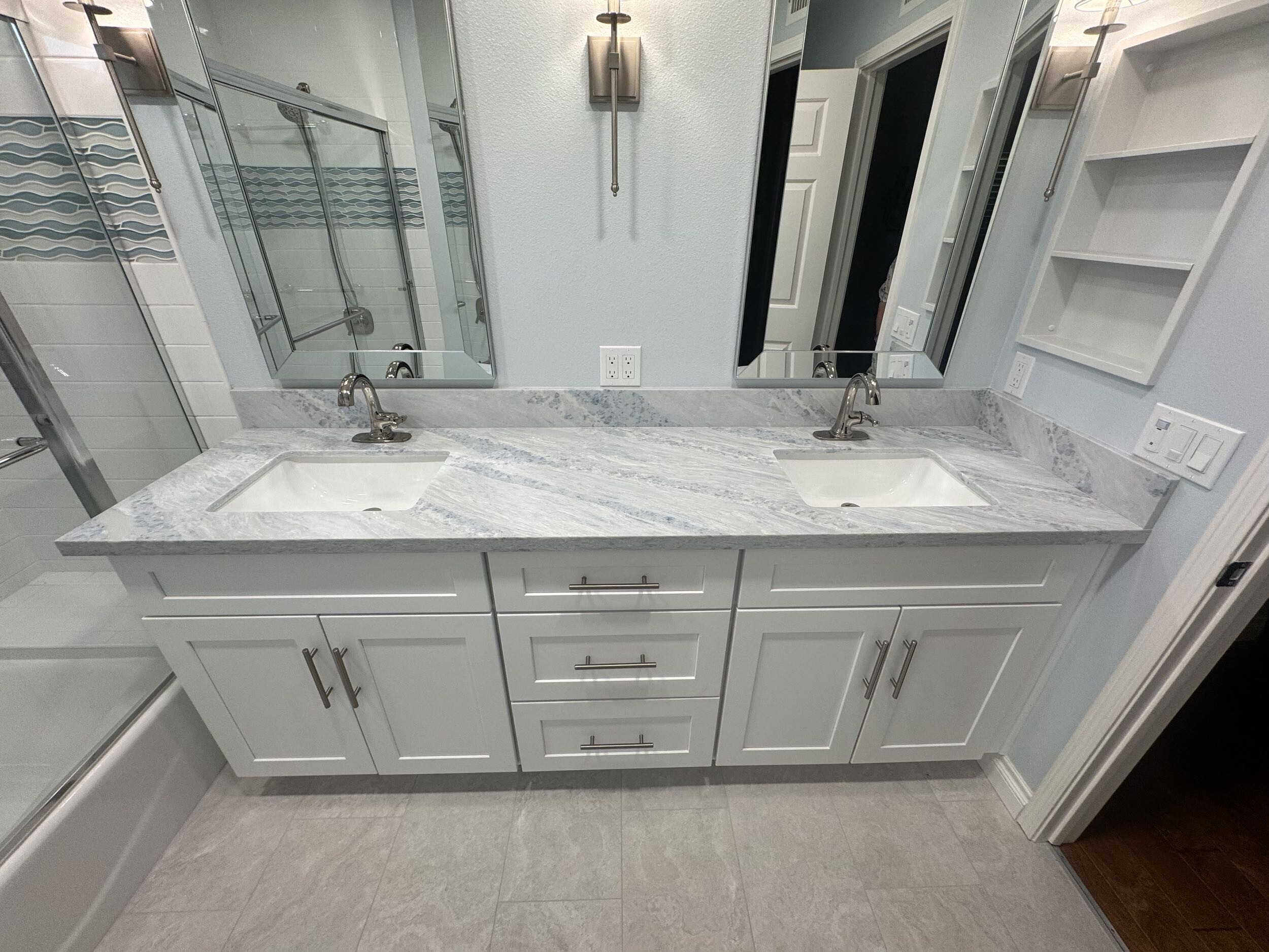 Modern white kitchen with island seating, stainless steel appliances, pendant lights, gray backsplash, wood floors, and bright natural light.
