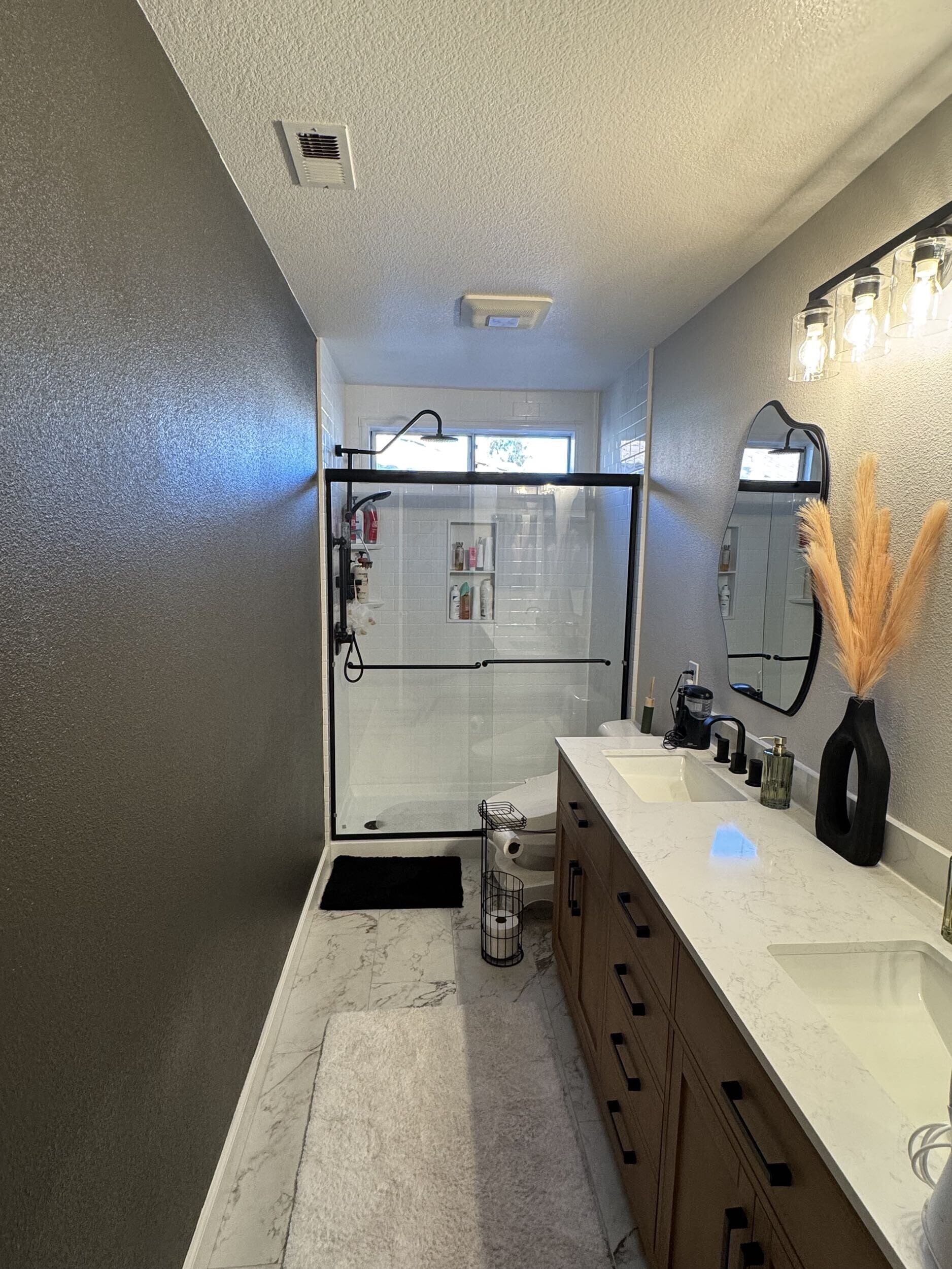 Modern bathroom remodel with double vanity, quartz countertop, black fixtures, framed glass shower enclosure, and marble-look flooring.