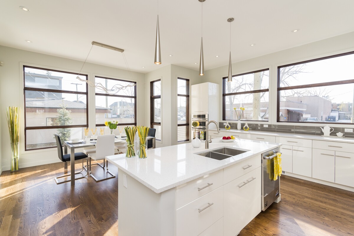 Modern kitchen remodel with white cabinetry, large quartz island, stainless steel sink, pendant lighting, hardwood floors, and dining area windows.