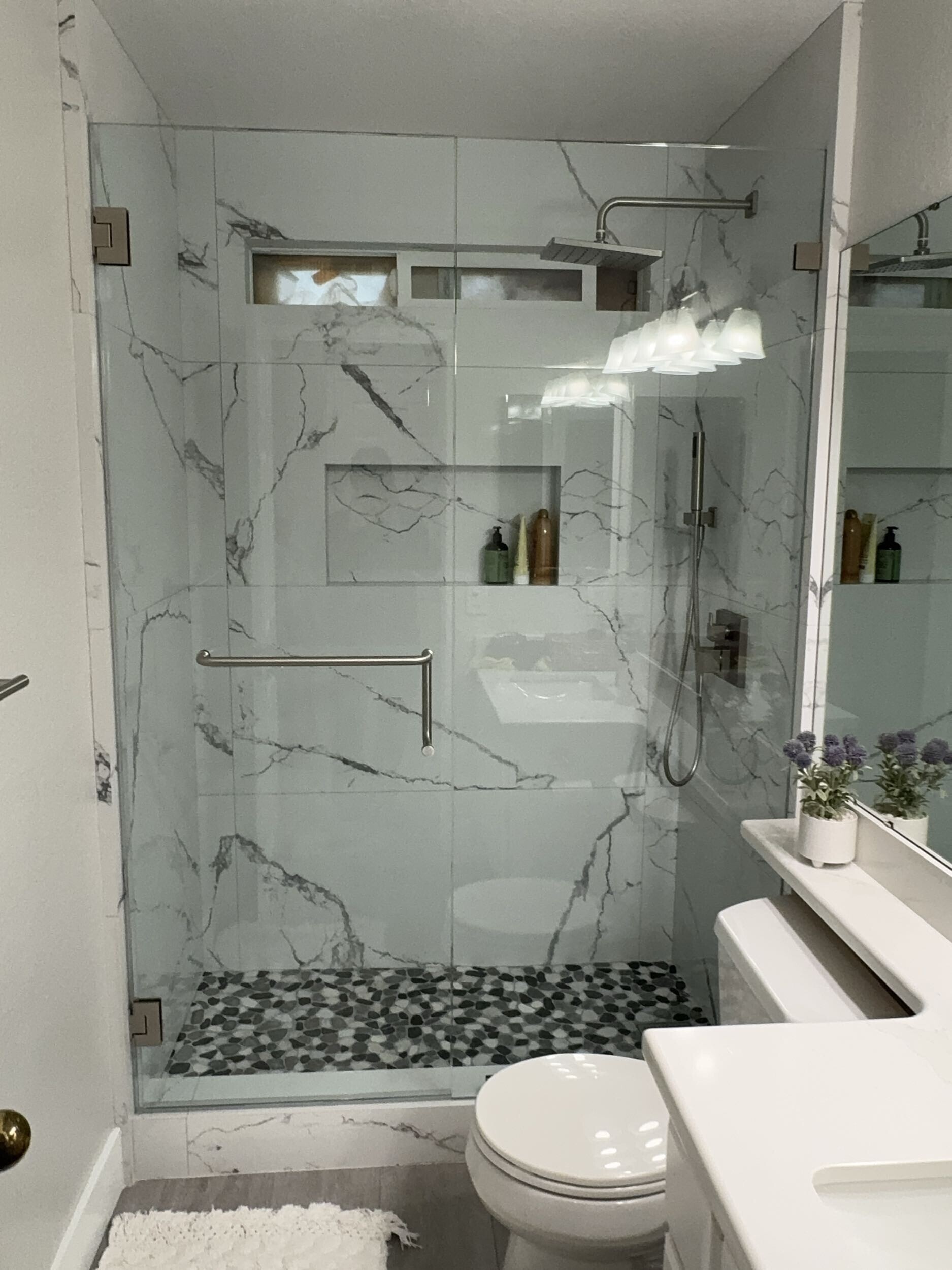 Modern bathroom remodel with frameless glass walk-in shower, marble-look wall tile, pebble shower floor, floating vanity, and sleek matte fixtures.