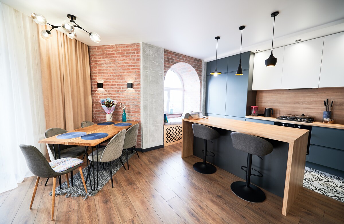 Contemporary open-concept kitchen and dining area with wood finishes, pendant lighting, brick accent wall, breakfast bar seating, and modern cabinetry.