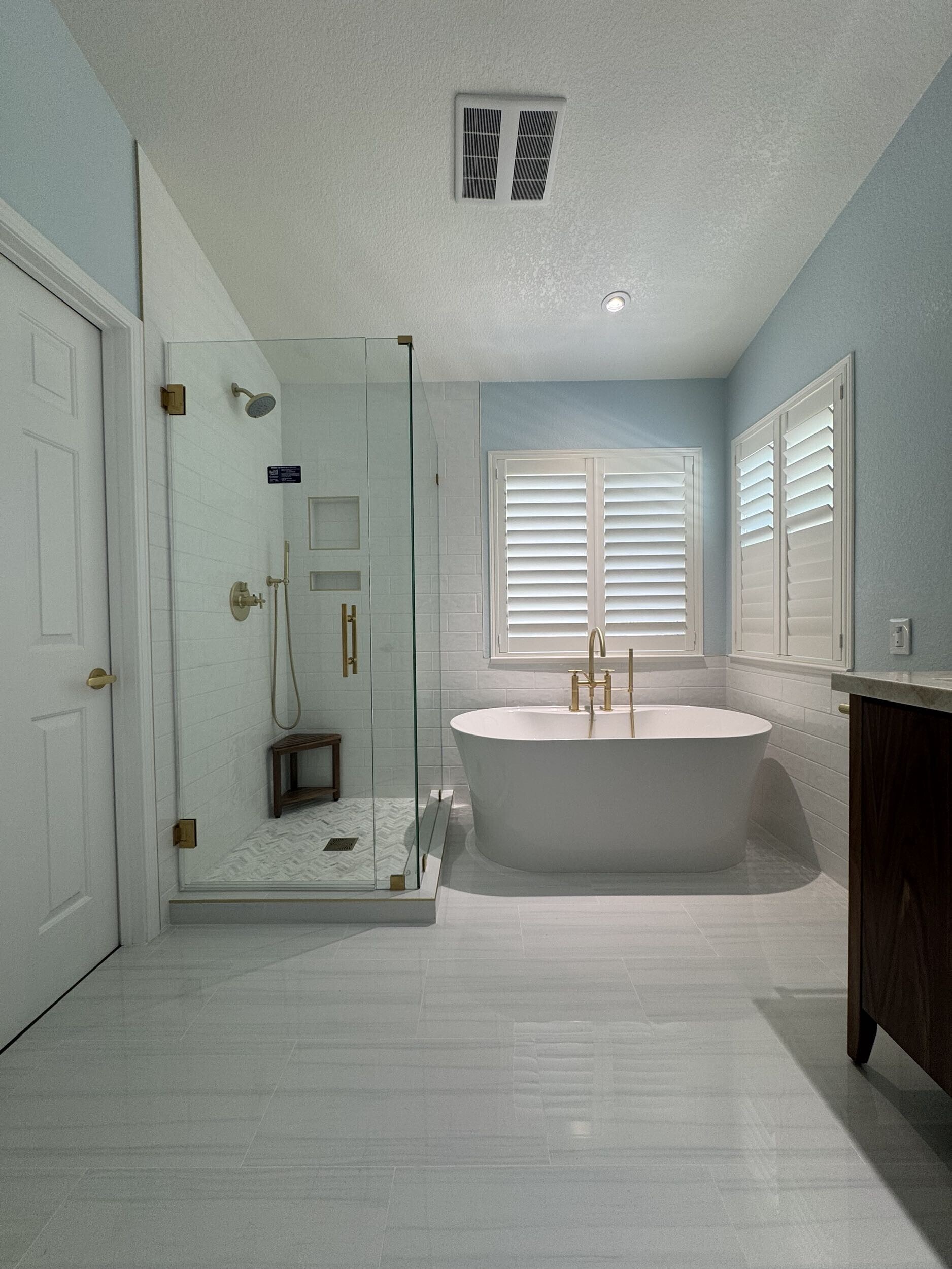 Luxury bathroom remodel with freestanding soaking tub, frameless glass shower, gold fixtures, white tile flooring, and bright modern finishes.
