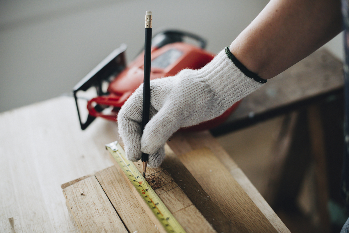 A gloved hand uses a pencil and measuring tape to carefully mark a piece of wood near a power tool, essential steps when planning home remodeling projects.