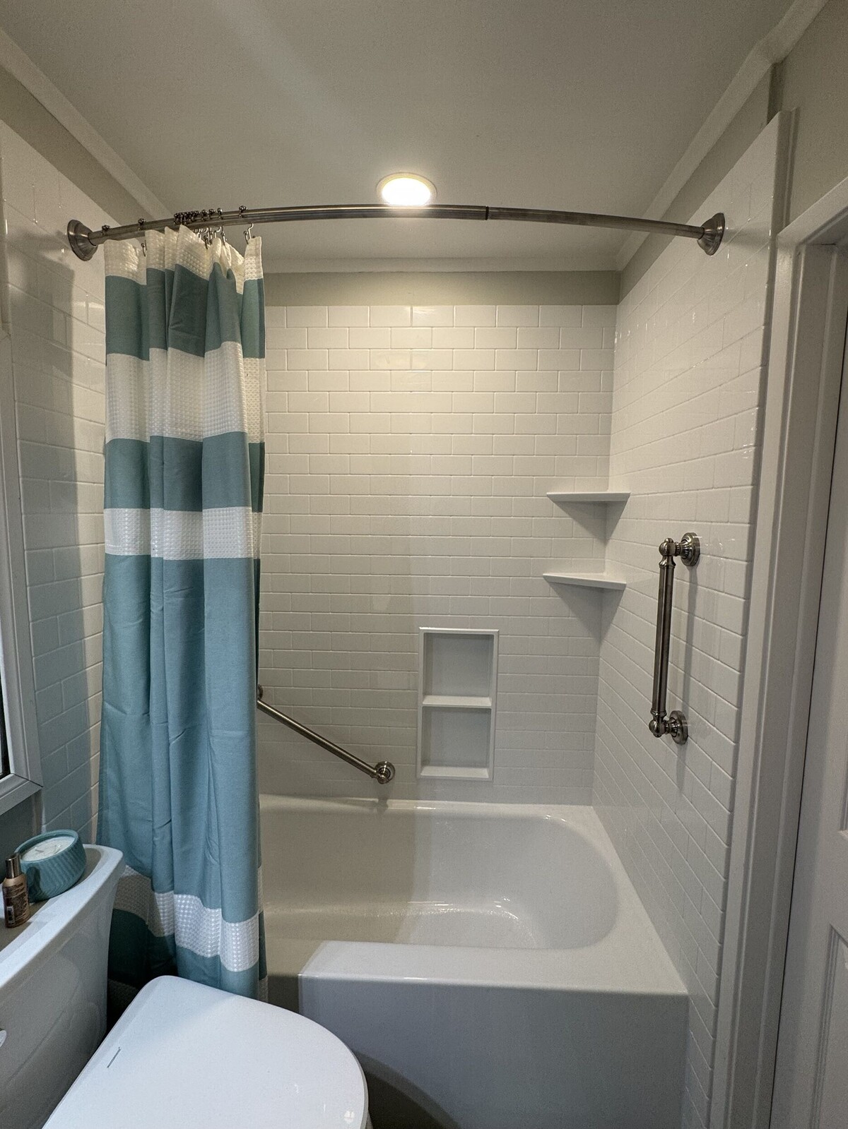 A clean bathroom features a white subway-tile acrylic shower-and-tub combo equipped with safety grab bars, integrated shelving, and a teal-and-white striped curtain.